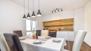 Zona pranzo moderna con tavolo bianco apparecchiato per quattro persone, lampade a sospensione nere e una cucina sullo sfondo con mobili in legno e lavello in acciaio inossidabile.