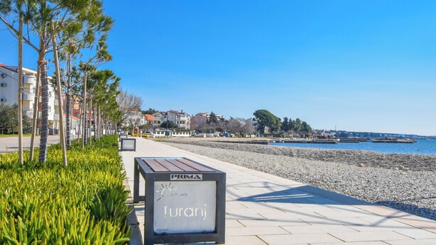 Una passeggiata lungomare con un cartello che indica 'Turanj' e vista sul mare e sugli edifici sullo sfondo.