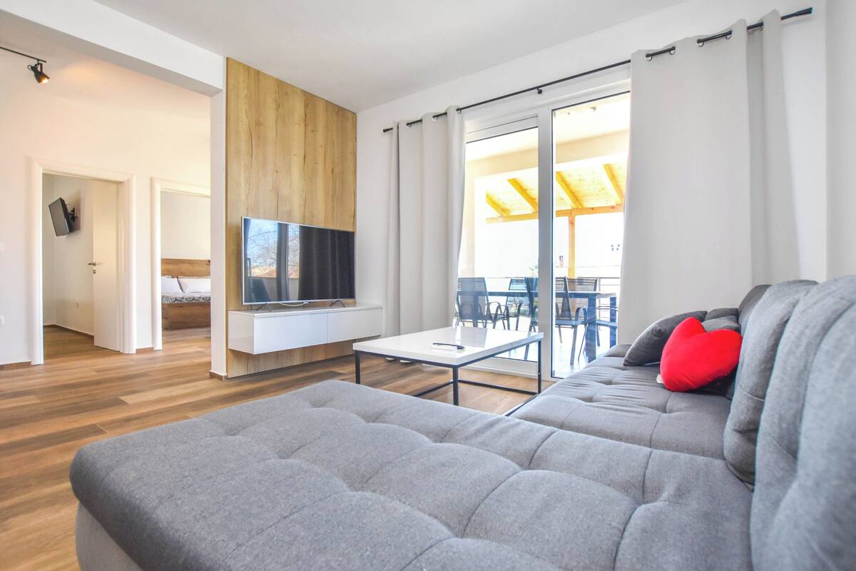Un salon moderne avec un canapé gris, un téléviseur à écran plat fixé sur un panneau en bois, et un coin repas comprenant une table et des chaises.