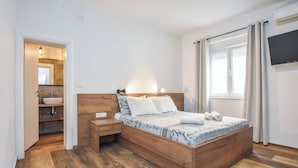 Una camera da letto con un letto di legno, un comodino, una mensola portaoggetti e una TV a parete, oltre a una finestra con tende.