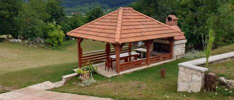 A wooden gazebo with a tiled roof, stone walls, and a stone fireplace.