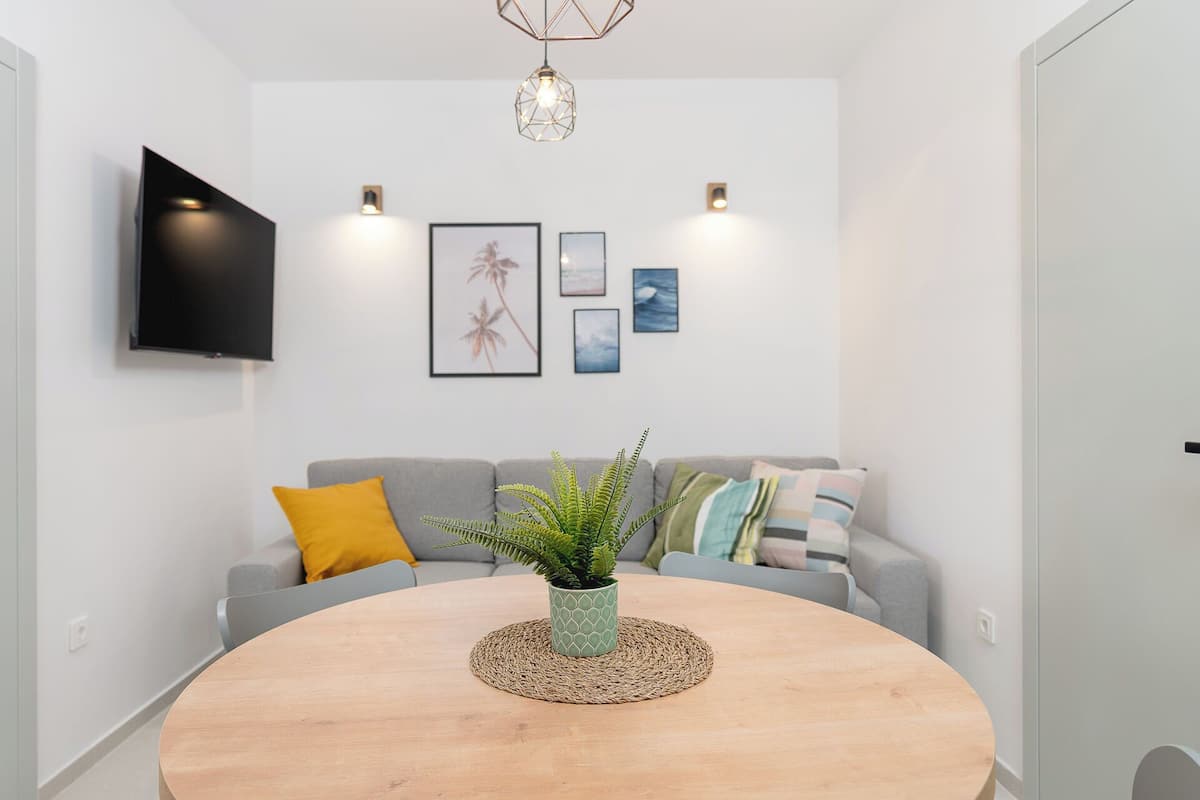 Een moderne woonkamer met een ronde houten tafel, grijze banken, een wandgemonteerde televisie en wandlampen.