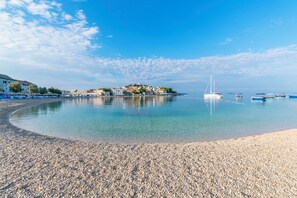 A pebbled beach with clear water, boats, and buildings in the background. - Apartment Conte Cikara (Primosten)