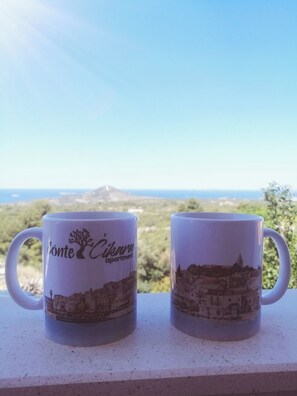 Two white mugs with a landscape design, placed on a ledge with a view of a coastal area and a mountain in the background. - Apartment Conte Cikara (Primosten)