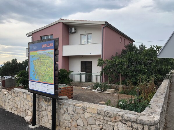 A two-story building with a pink facade, a balcony, and a stone wall in front. - Apartment Conte Cikara (Primosten)