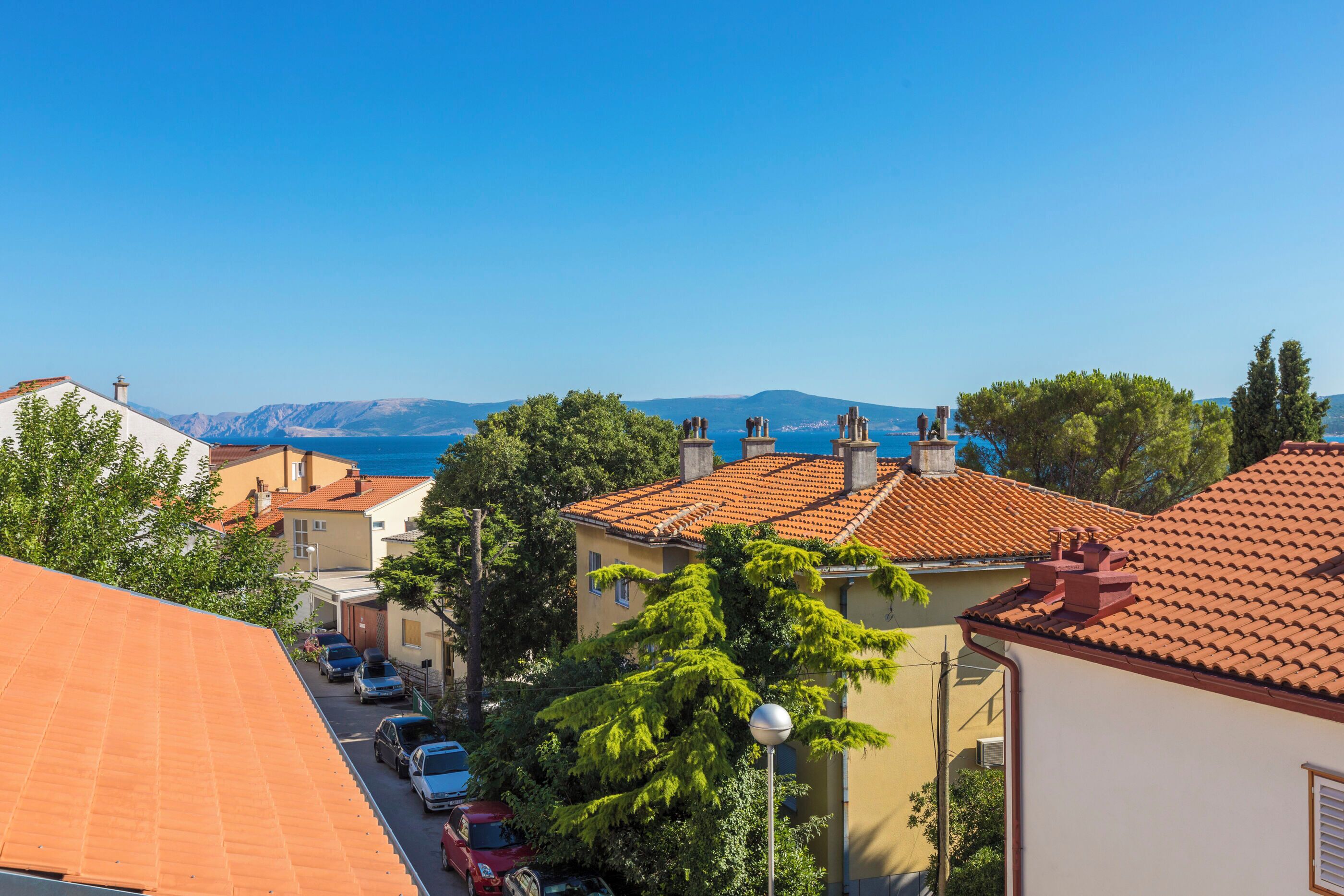 Un barrio residencial con techos de tejas de arcilla, coches aparcados y una vista despejada del mar y las montañas al fondo.