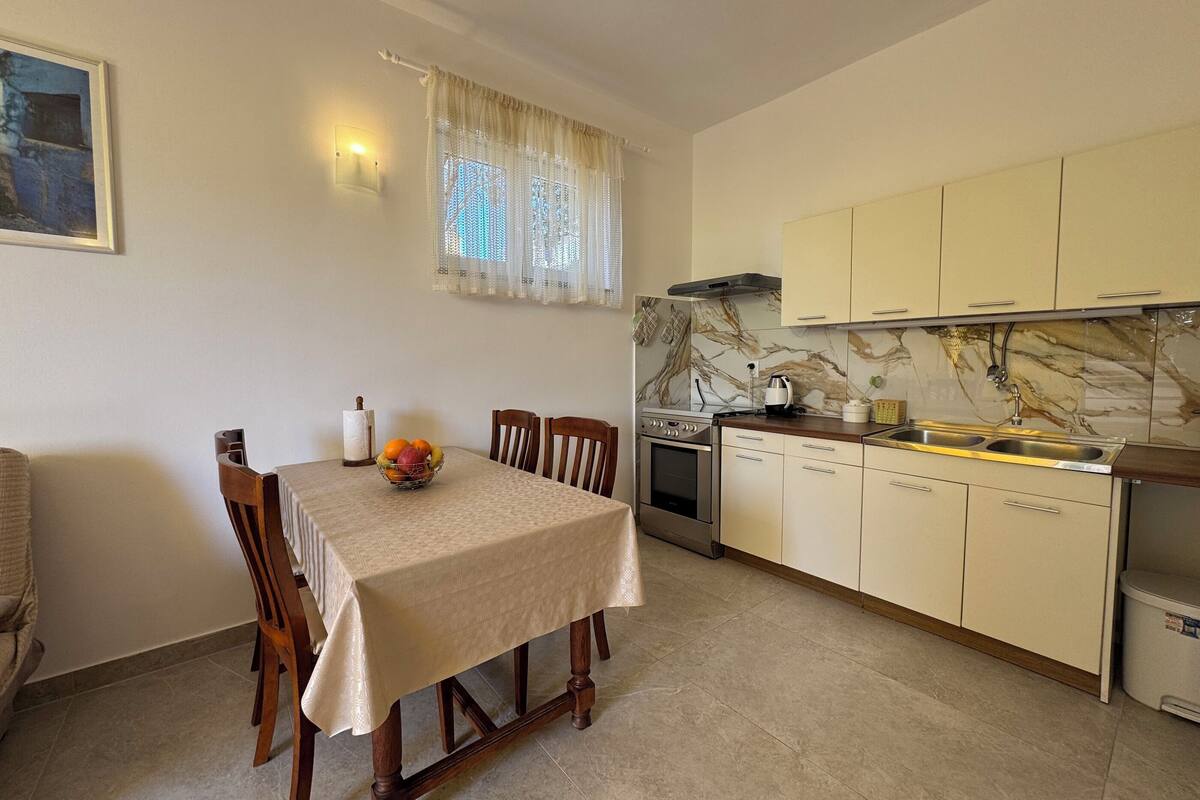 A dining area with a table, chairs, a fruit bowl, a wall-mounted light, and a kitchen with cabinets and a stove.