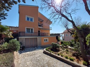 Ein zweistöckiges Gebäude mit Balkon, Steinweg und umgebender Bepflanzung.