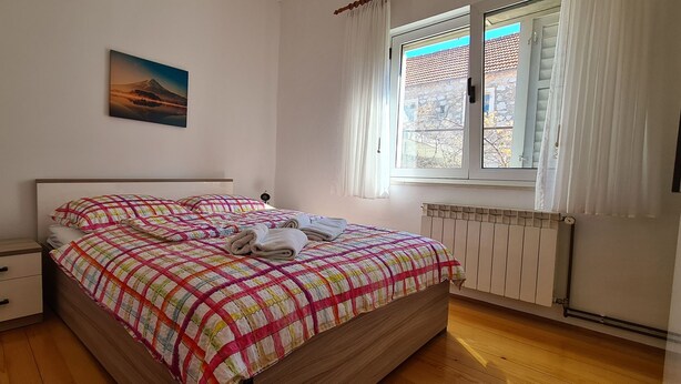 A bedroom with a bed, a nightstand, a wall-mounted picture, a window with curtains, and a radiator. - Apartment Turan (Jelsa)