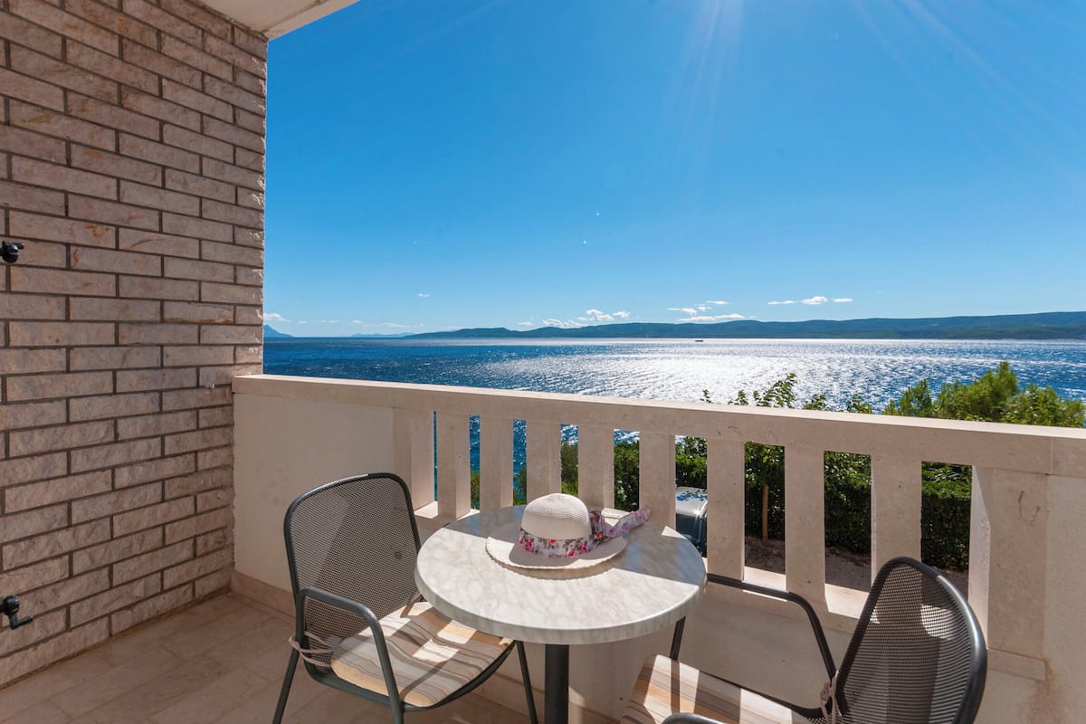 A balcony with a view of a body of water and distant mountains. There is a table with a hat and a chair.