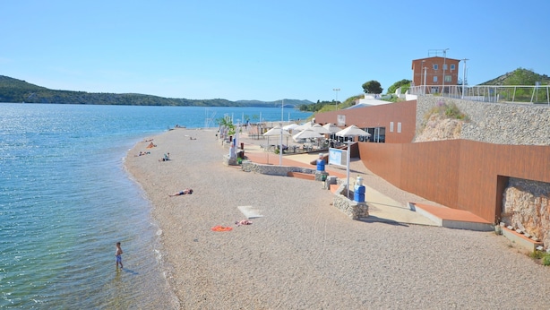 A beach with a pebble shore, umbrellas, and a building with a red roof. - Apartment Kata (Sibenik)