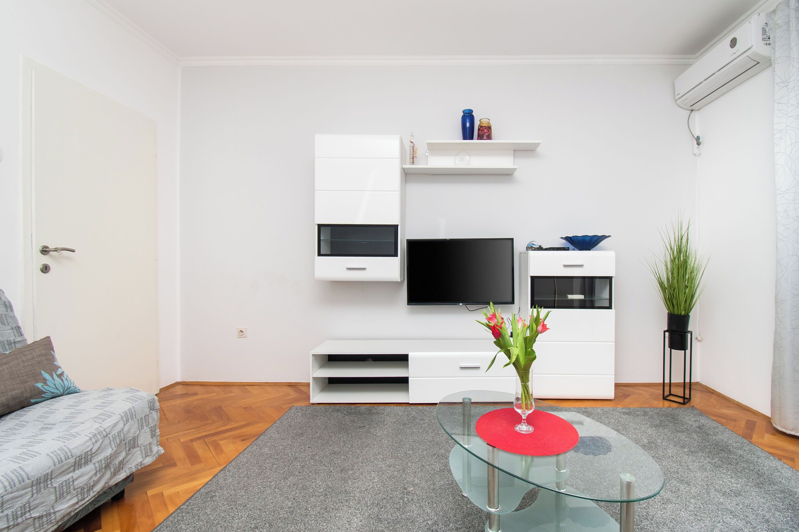 Ein modernes Wohnzimmer mit einem Flachbildfernseher, weißen Schränken, einem Glastisch und einer Topfpflanze.