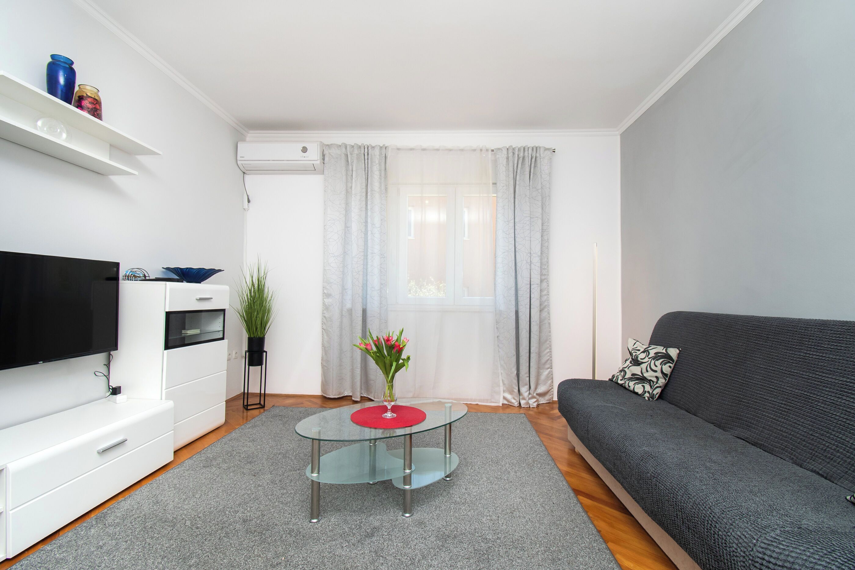 A modern living room with a flat-screen TV, a glass coffee table, a grey sofa, and a small side table with a vase of flowers.