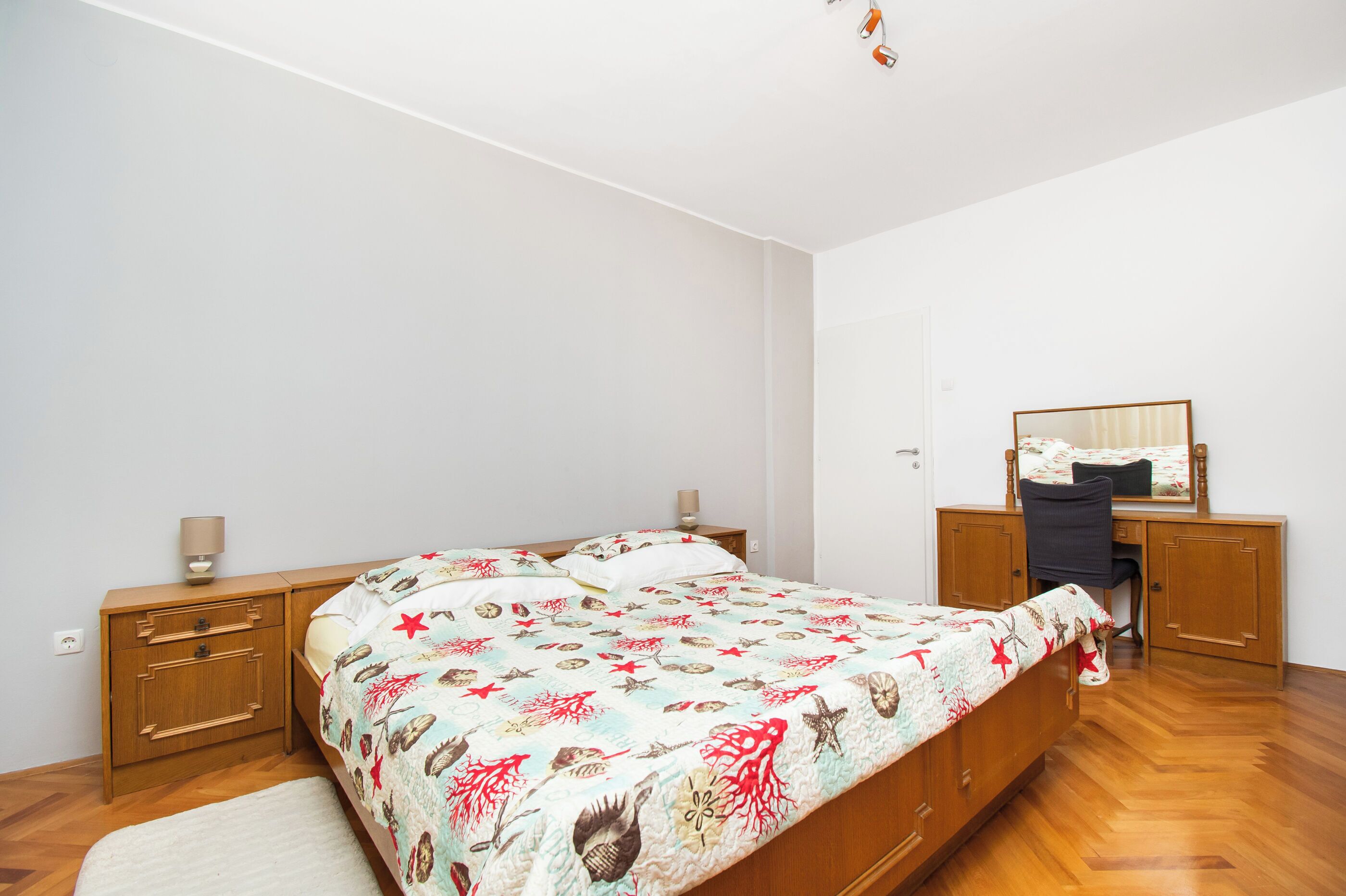 A bedroom with a bed, wooden nightstands, a desk with a chair, and a mirror.