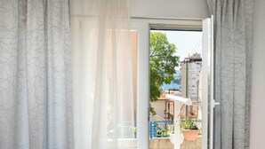 A room with a large window, white curtains, and a view of a balcony with potted plants and a blue railing. - Apartment Kata (Sibenik)