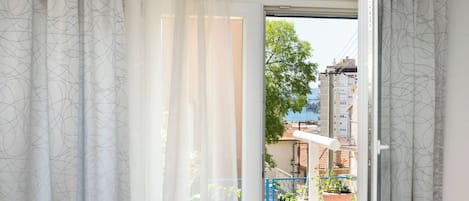 A room with a large window, white curtains, and a view of a balcony with potted plants and a blue railing.