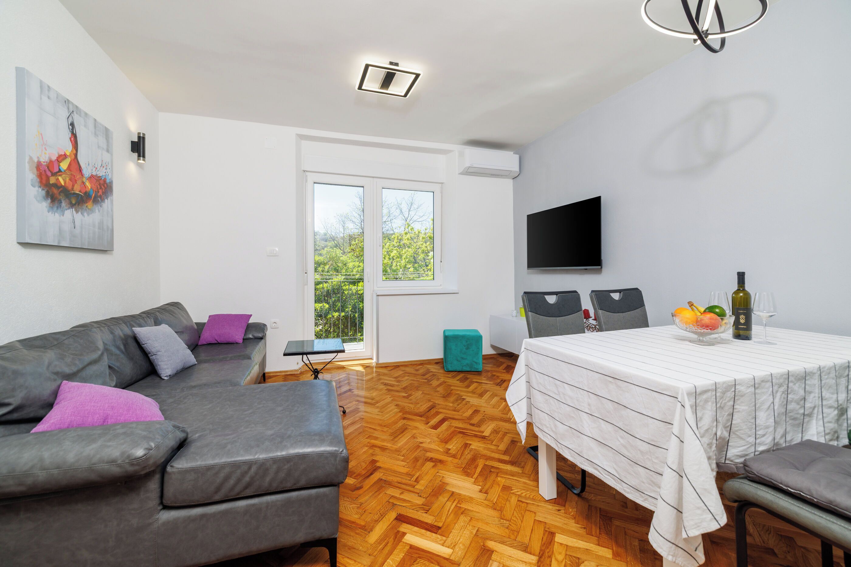 Ein modernes Wohnzimmer mit einem grauen Sofa, einem Esstisch mit weißer Tischdecke und einem an der Wand montierten Fernseher.