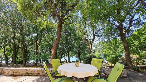 Une table ronde recouverte d’une nappe blanche, entourée de quatre chaises vertes, est placée sur une terrasse en pierre, avec des arbres en arrière-plan.