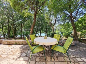 Une table ronde recouverte d’une nappe blanche, entourée de quatre chaises vertes, est placée sur une terrasse en pierre, avec des arbres en arrière-plan.