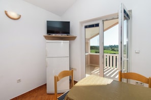 A small room with a table and chairs, a TV on a shelf, a refrigerator, and a sliding glass door leading to a balcony. - Apartments Nikola (Nin)