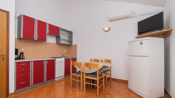 A compact kitchen with red cabinets, a white refrigerator, and a dining table with chairs. - Apartments Nikola (Nin)