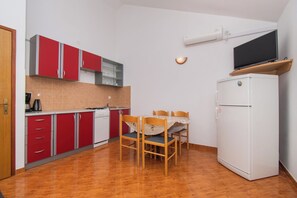 A compact kitchen with red cabinets, a white refrigerator, and a dining table with chairs. - Apartments Nikola (Nin)