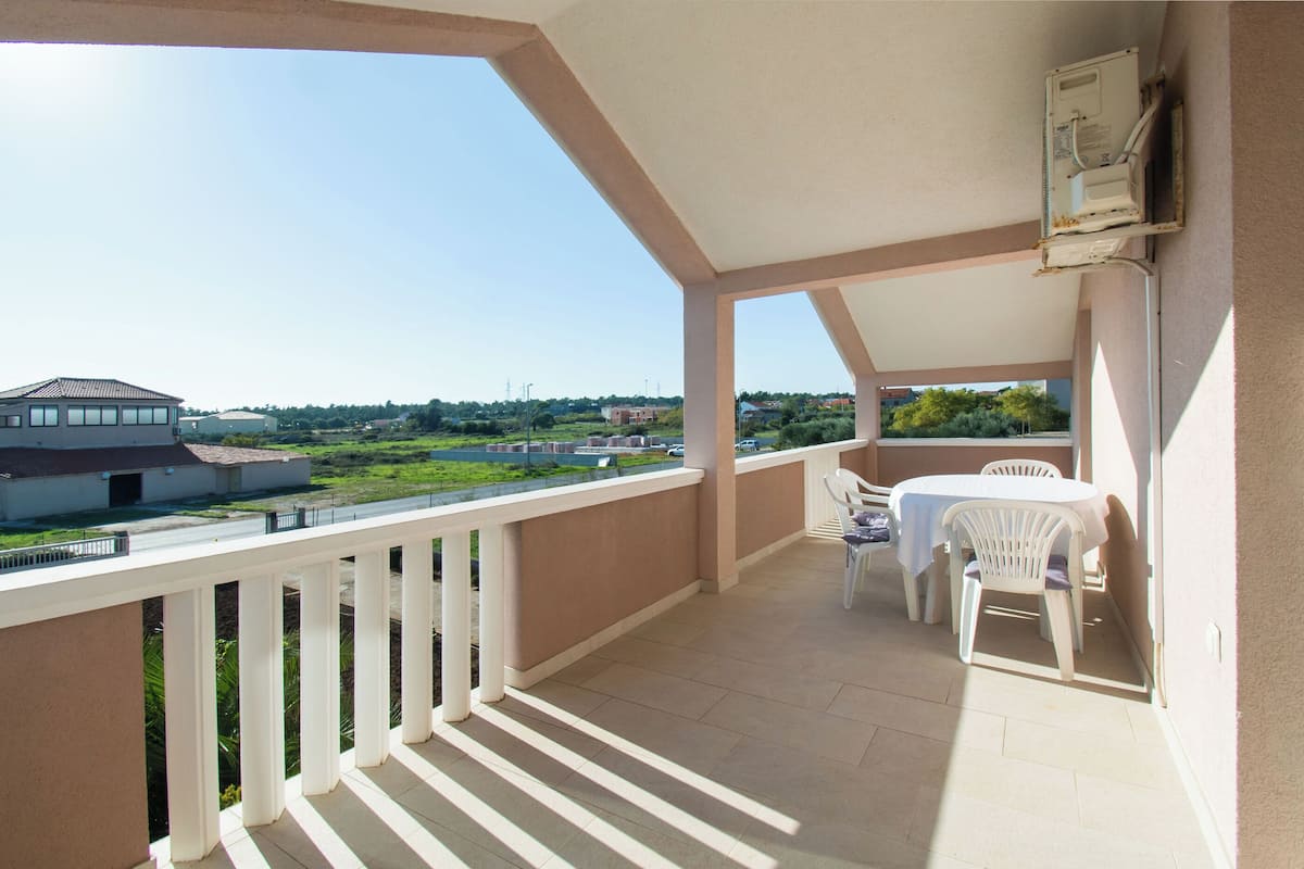 A balcony with a white railing, a table with chairs, and a view of a building and greenery.