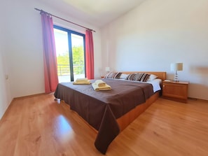 A bedroom with a large bed, wooden floor, a nightstand with a lamp, and a window with curtains. - Apartments Zelena Oaza (Novi Vinodolski)