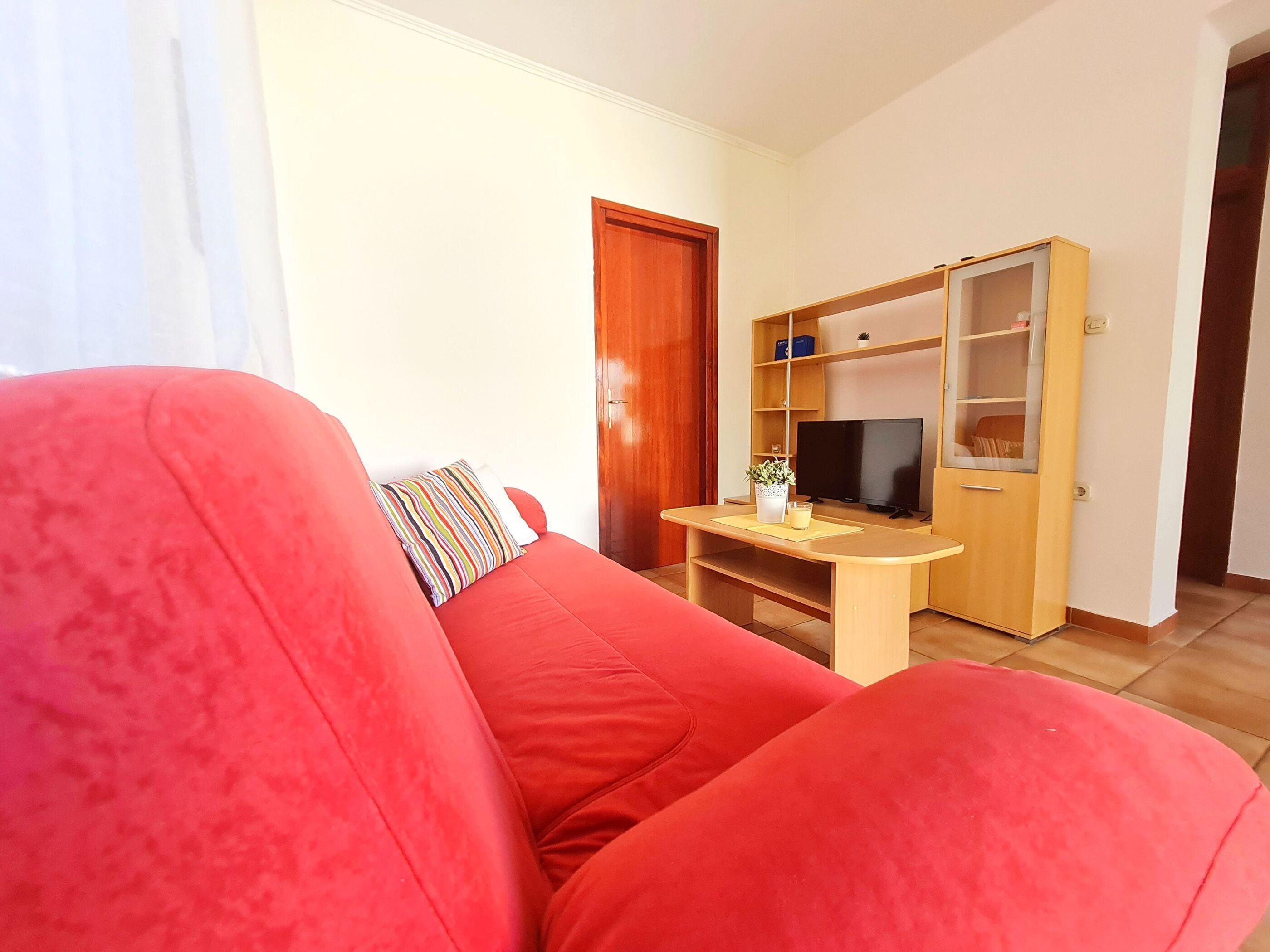 Un divano rosso con un cuscino a righe, un mobile TV in legno con televisore e un armadietto in legno con ripiani.