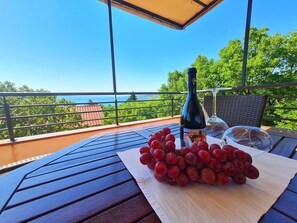 A table with a bottle of wine, a glass, and a bunch of grapes on a wooden table with a view of the sea and trees. - Apartments Zelena Oaza (Novi Vinodolski)