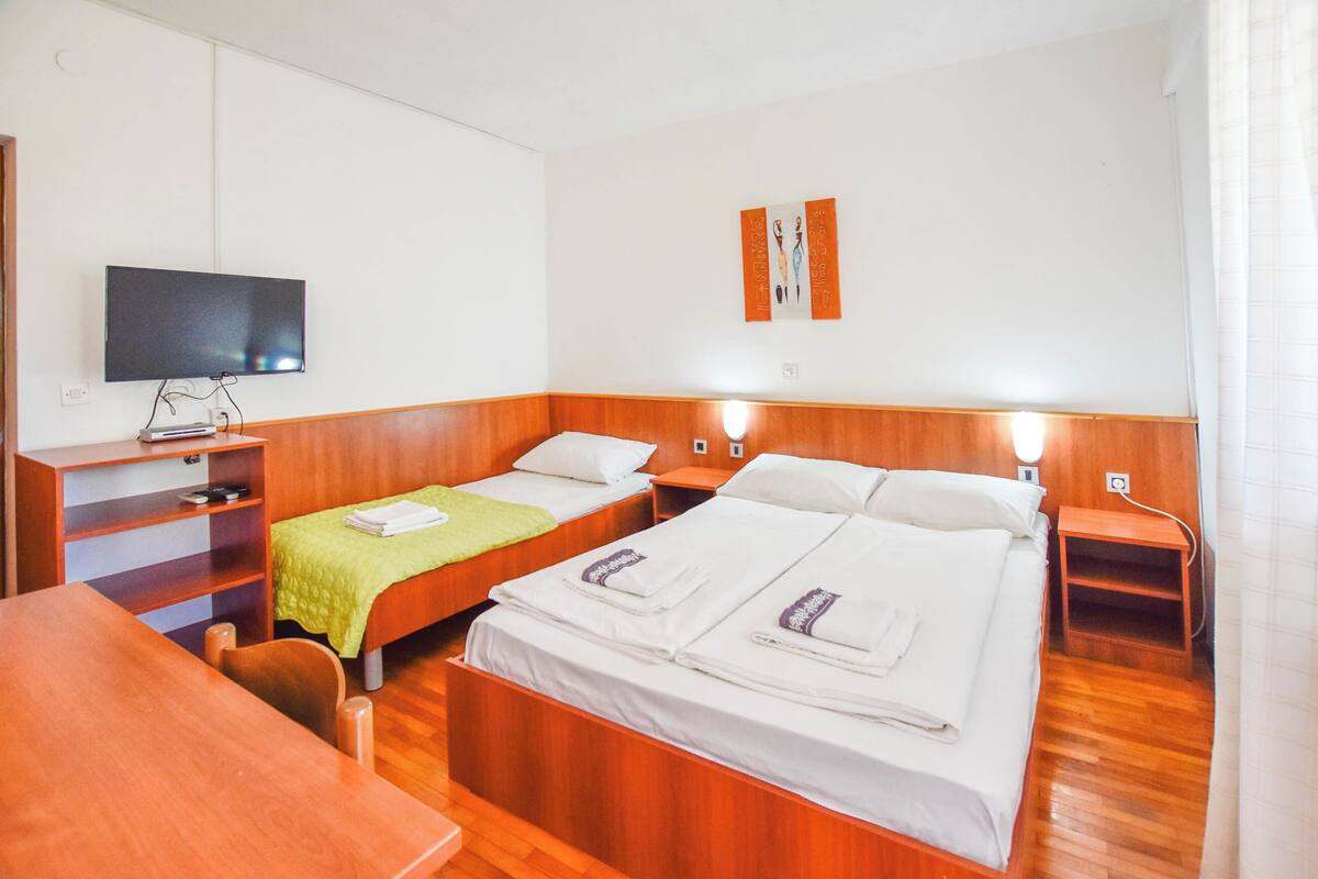 A hotel room with two single beds, a wooden desk, a television, and a small shelf with books.