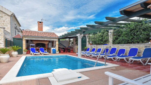 A pool area with blue lounge chairs, a covered seating area, and a stone building in the background. - Apartments Jacqueline (Pakostane)