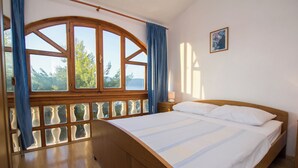 Ein Schlafzimmer mit einem hölzernen Bett, einem großen Fenster mit Vorhängen, einem Nachttisch und einem gerahmten Bild an der Wand.