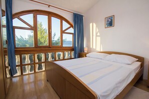 Ein Schlafzimmer mit einem hölzernen Bett, einem großen Fenster mit Vorhängen, einem Nachttisch und einem gerahmten Bild an der Wand.