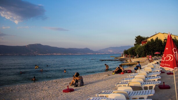 Ein Strand mit badenden und sonnenden Menschen, weißen Liegestühlen und einem roten Sonnenschirm mit dem Schriftzug 'GIDA'.
