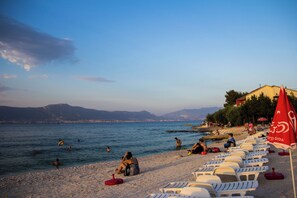 Una playa con gente nadando y tomando el sol, sillas de descanso blancas y un paraguas rojo con la palabra 'GIDA'.