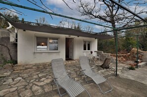 A small, single-story building with a stone exterior and a covered patio area. There are two lounge chairs and a table on the patio. - House Roman (Rijeka)