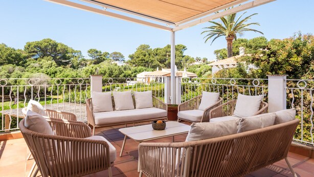A covered outdoor seating area with wicker furniture and a view of greenery and buildings in the background. - Villa Maite by Escape Home (Binibequer)
