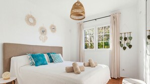 A neatly made bed with a white duvet and rolled towels, a wooden headboard, and a window with curtains. - Villa Maite by Escape Home (Binibequer)