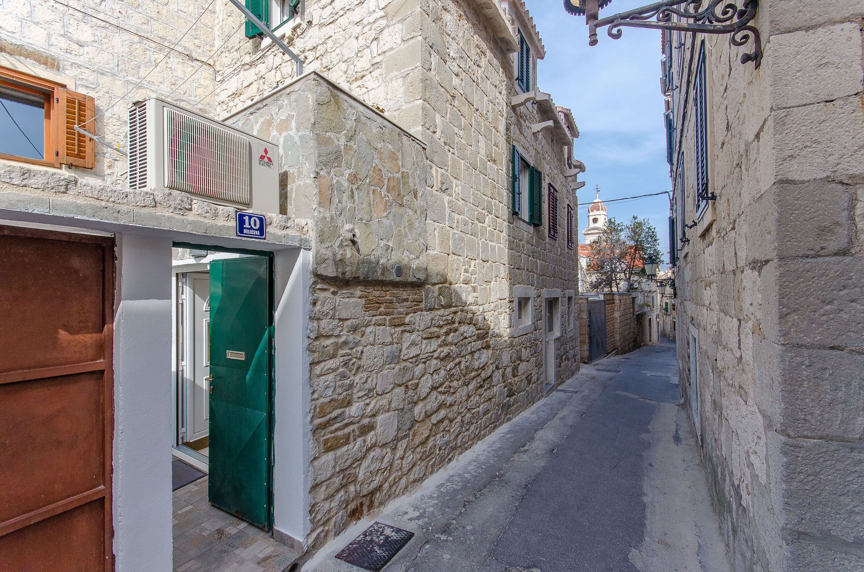 Eine enge Gasse mit Steinbauten, einer gut sichtbaren Tür mit einer Hausnummer und einem klaren Himmel.