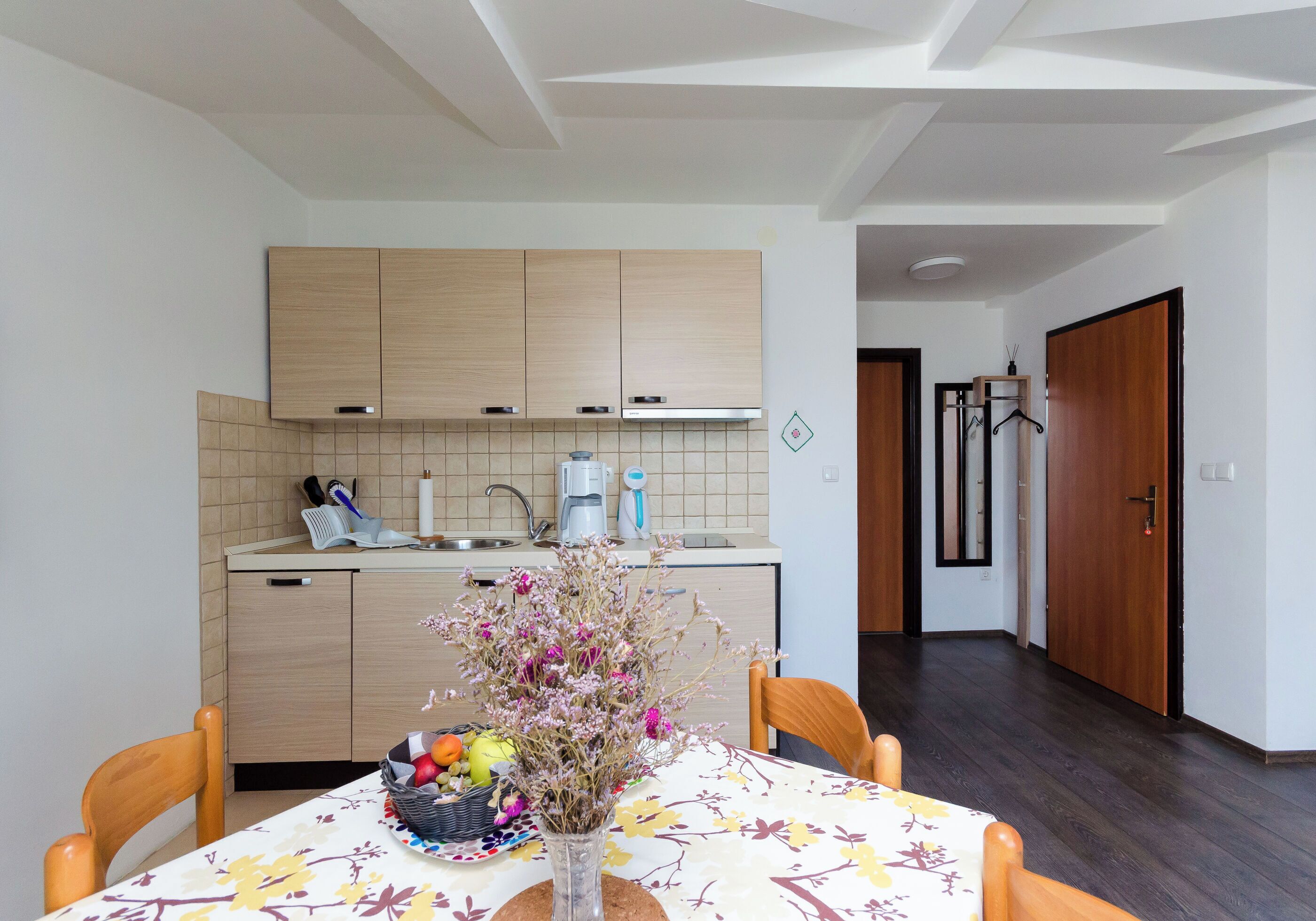 A modern kitchen with wooden cabinets, a dining table with a floral tablecloth, and a vase with flowers.