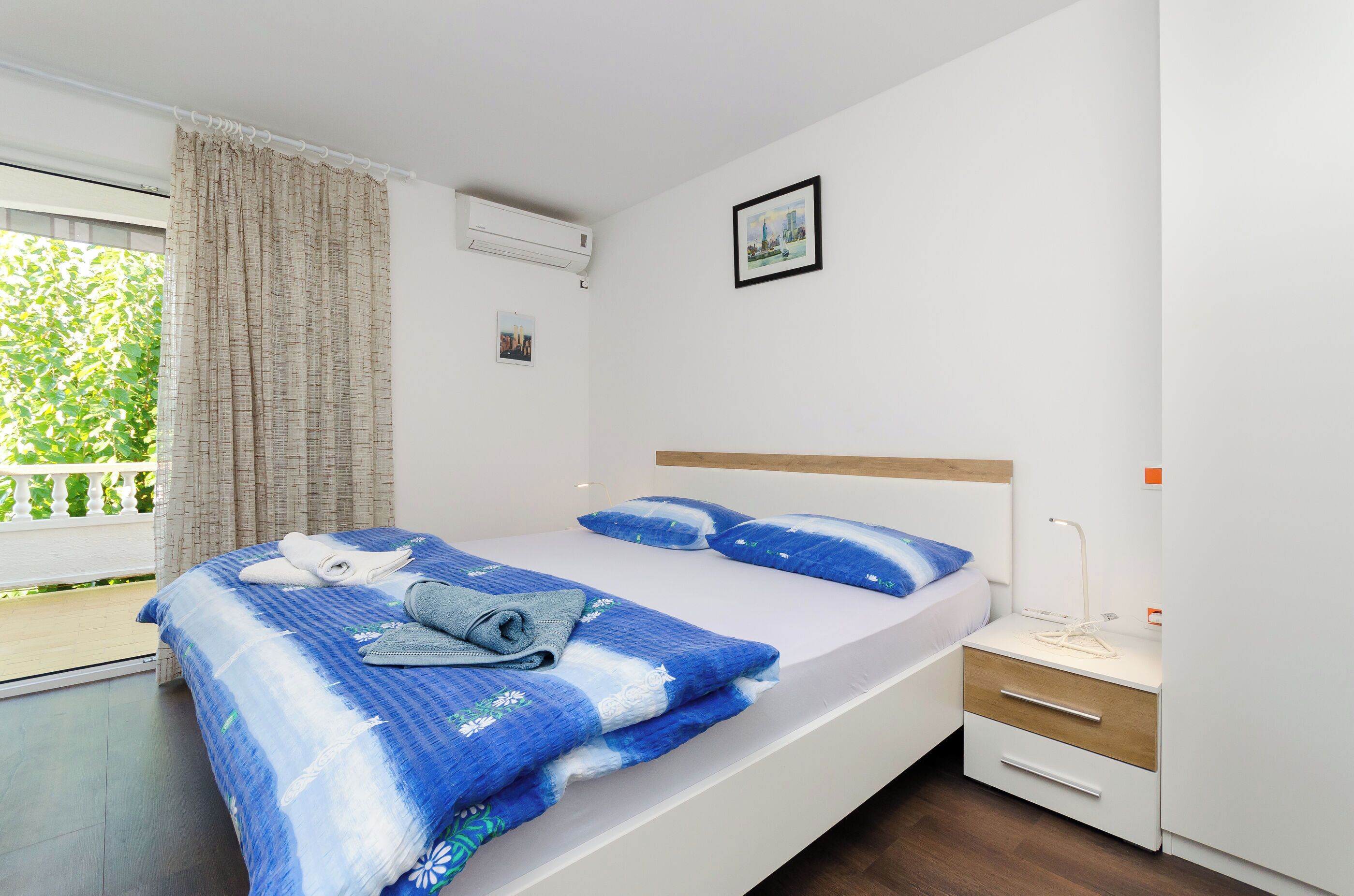 A neatly made bed with blue bedding, a bedside table with a lamp, and a window with curtains.