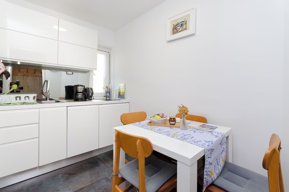 Cocina compacta con armarios blancos, una mesa de comedor con mantel floral y un pequeño rincón de estar con sillas de madera.
