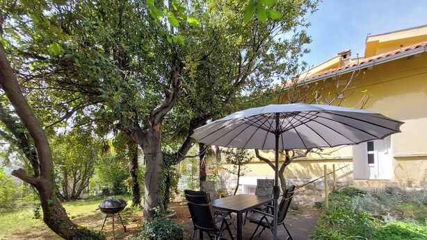 An outdoor patio with a table and chairs under a large umbrella, surrounded by trees and greenery. - Apartments Ineska (Crikvenica)