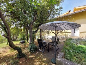 Patio esterno con tavolo e sedie sotto un ampio ombrellone, circondato da alberi e vegetazione.
