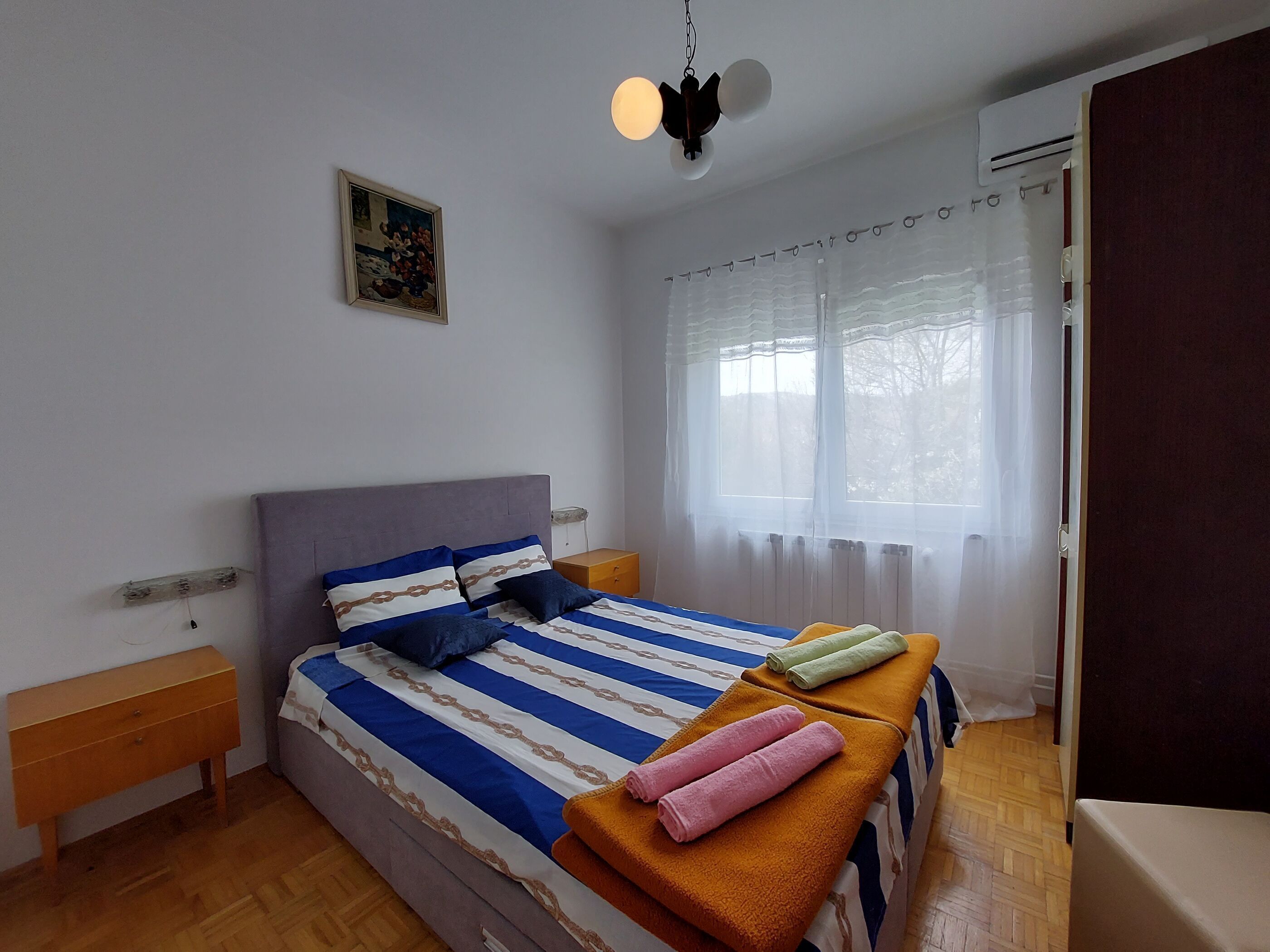 Een slaapkamer met een bed, nachtkastje, houten dressoir, schilderij aan de muur en een raam met dunne gordijnen.
