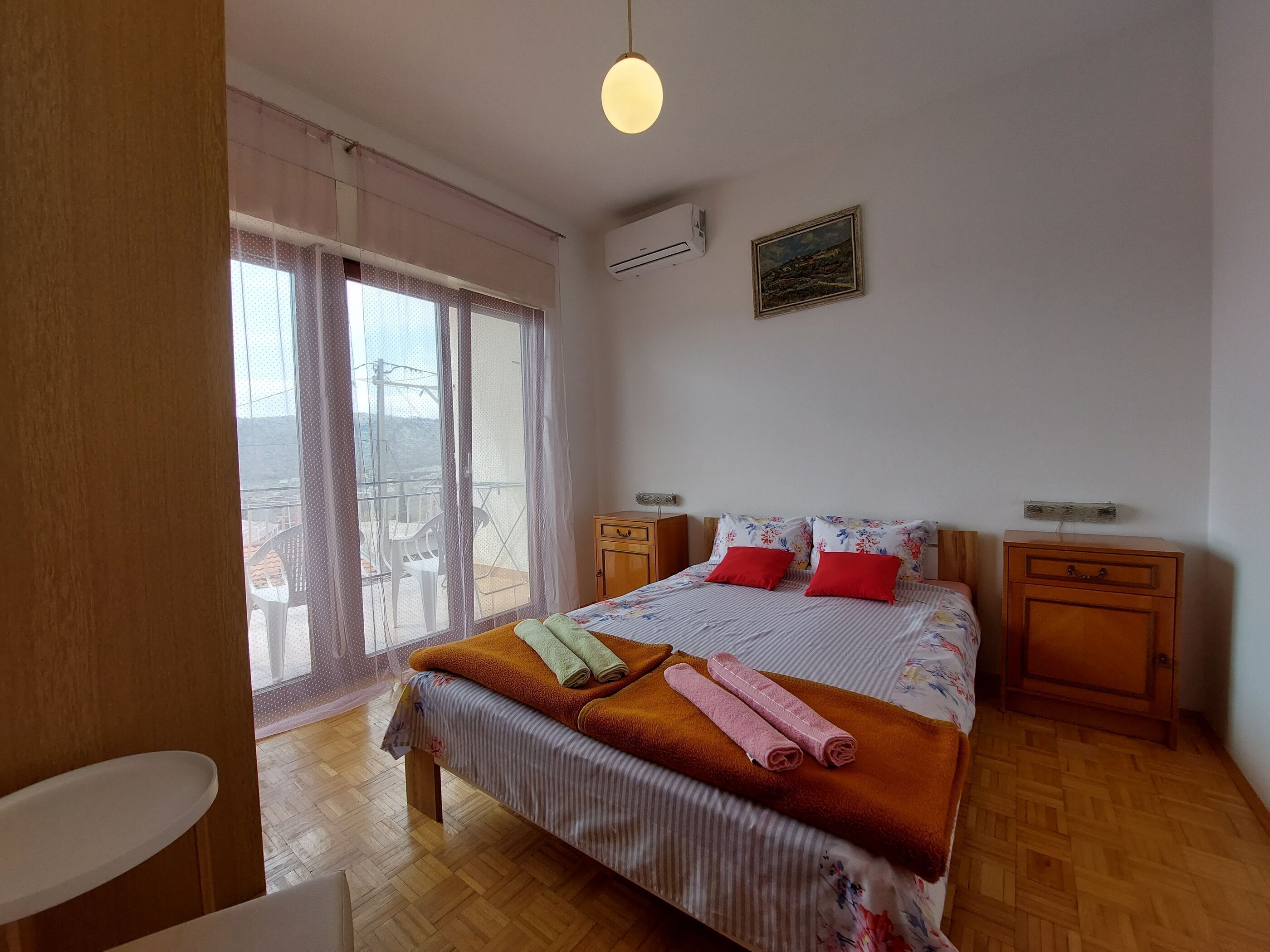 Ein Schlafzimmer mit einem Bett, zwei Nachttischen, Holzboden, einem Fenster mit Vorhängen und einer Klimaanlage.