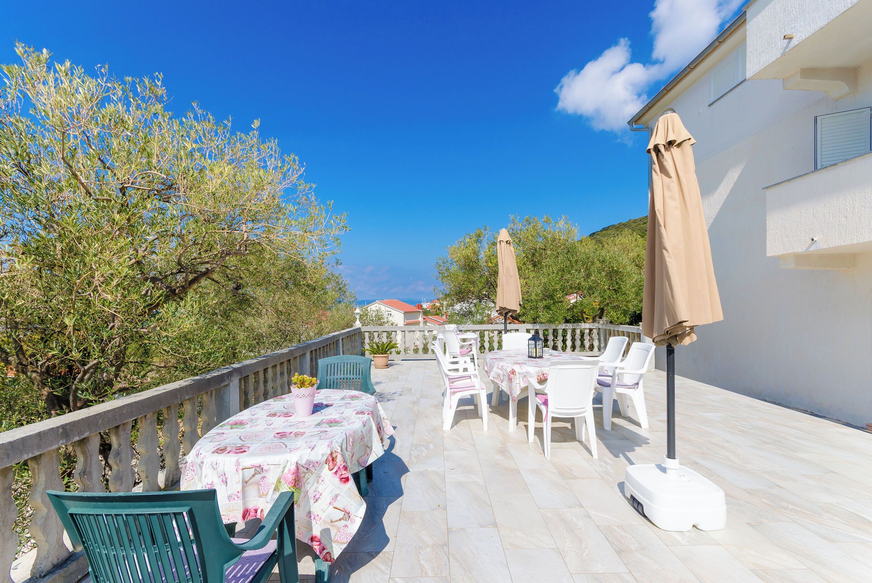 An outdoor patio with a table set for two, surrounded by white chairs and a beige umbrella.