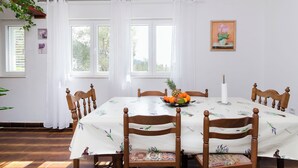 Un coin repas avec une table dressée pour quatre personnes, entourée de chaises en bois, d’une nappe blanche et d’un panier de fruits.