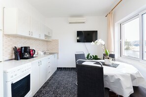 Een moderne keuken met witte kasten, een inbouwoven en een koffiezetapparaat. Er is een zithoek met een tafel en stoelen, en een televisie aan de muur bevestigd.
